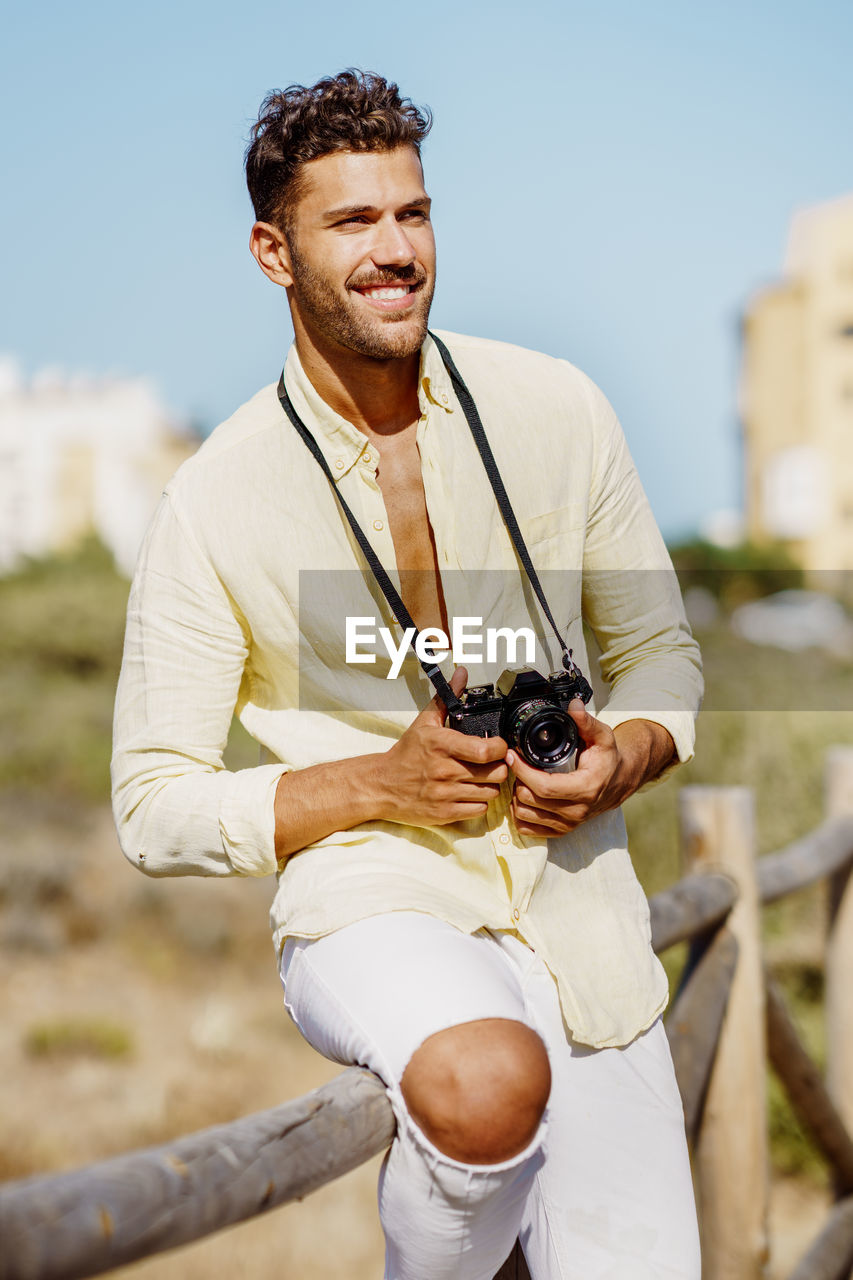 Smiling young man holding camera standing by railing against sky