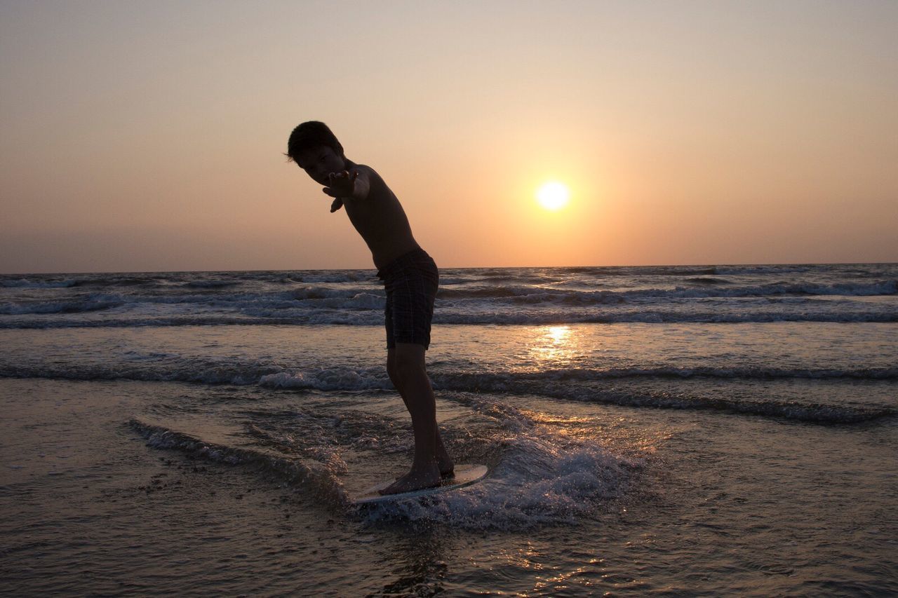 Shirtless boy surfing in sea against sky during sunset