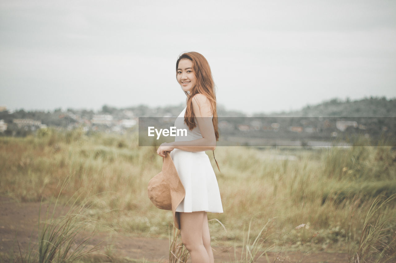 PORTRAIT OF HAPPY WOMAN STANDING ON GRASS AGAINST SKY