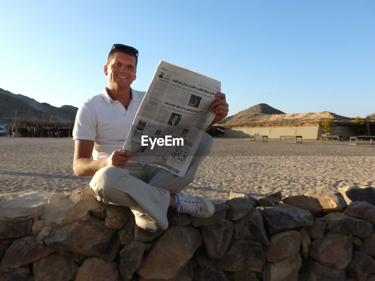 Full length portrait of smiling man with newspaper sitting on stone wall at beach