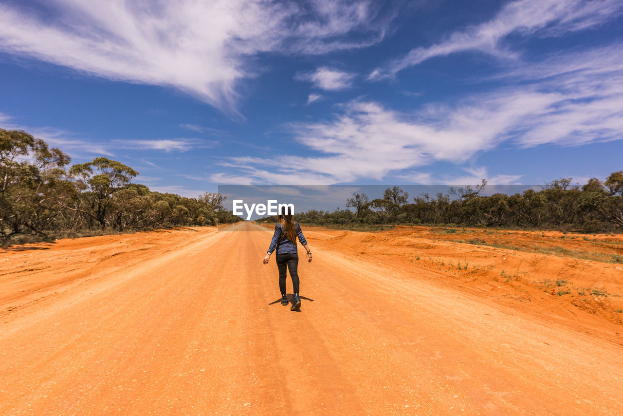 Rear view of woman walking on dirt road against sky