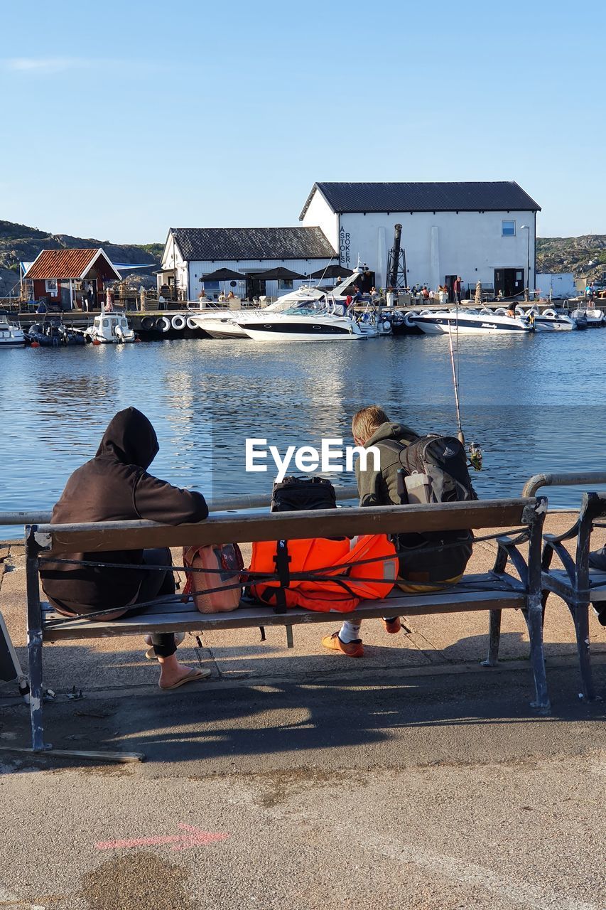 PEOPLE SITTING ON BOAT AT HARBOR BY BUILDINGS AGAINST SKY