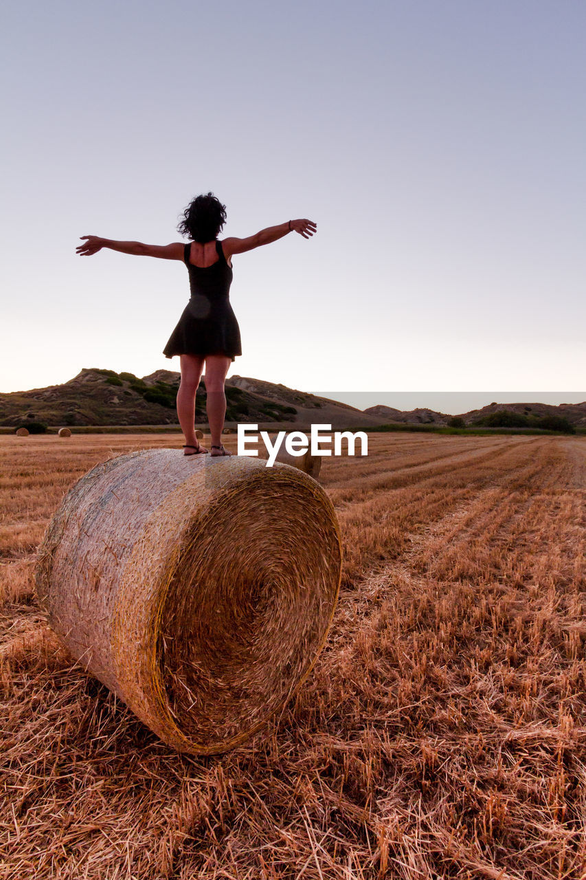 Caucasian 30s brunette woman, standing on an hay bale with raised arms