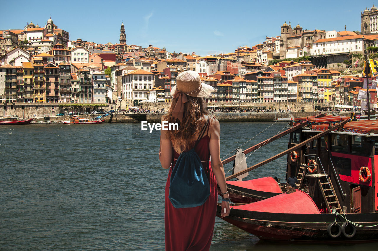 Rear view of young woman with backpack standing by river against sky in city