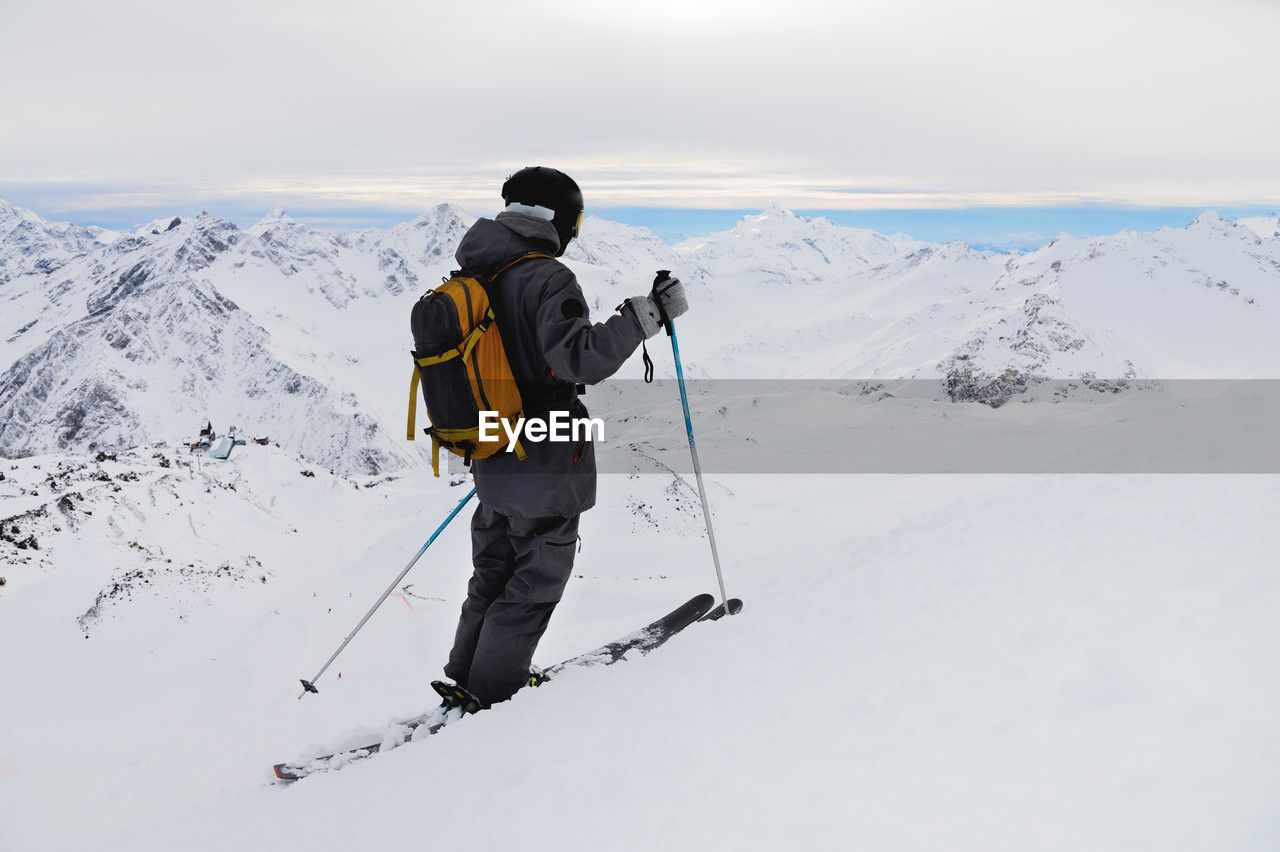 Side view of an athletic skier in a helmet and goggles with a backpack, standing on skis, holding