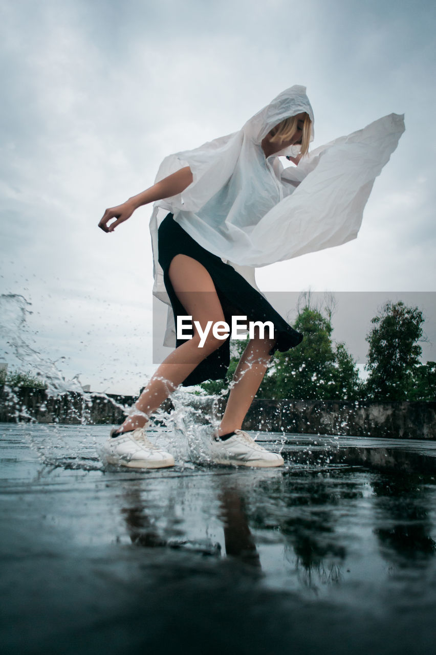 Full length of young woman wearing raincoat while jumping in puddle against sky