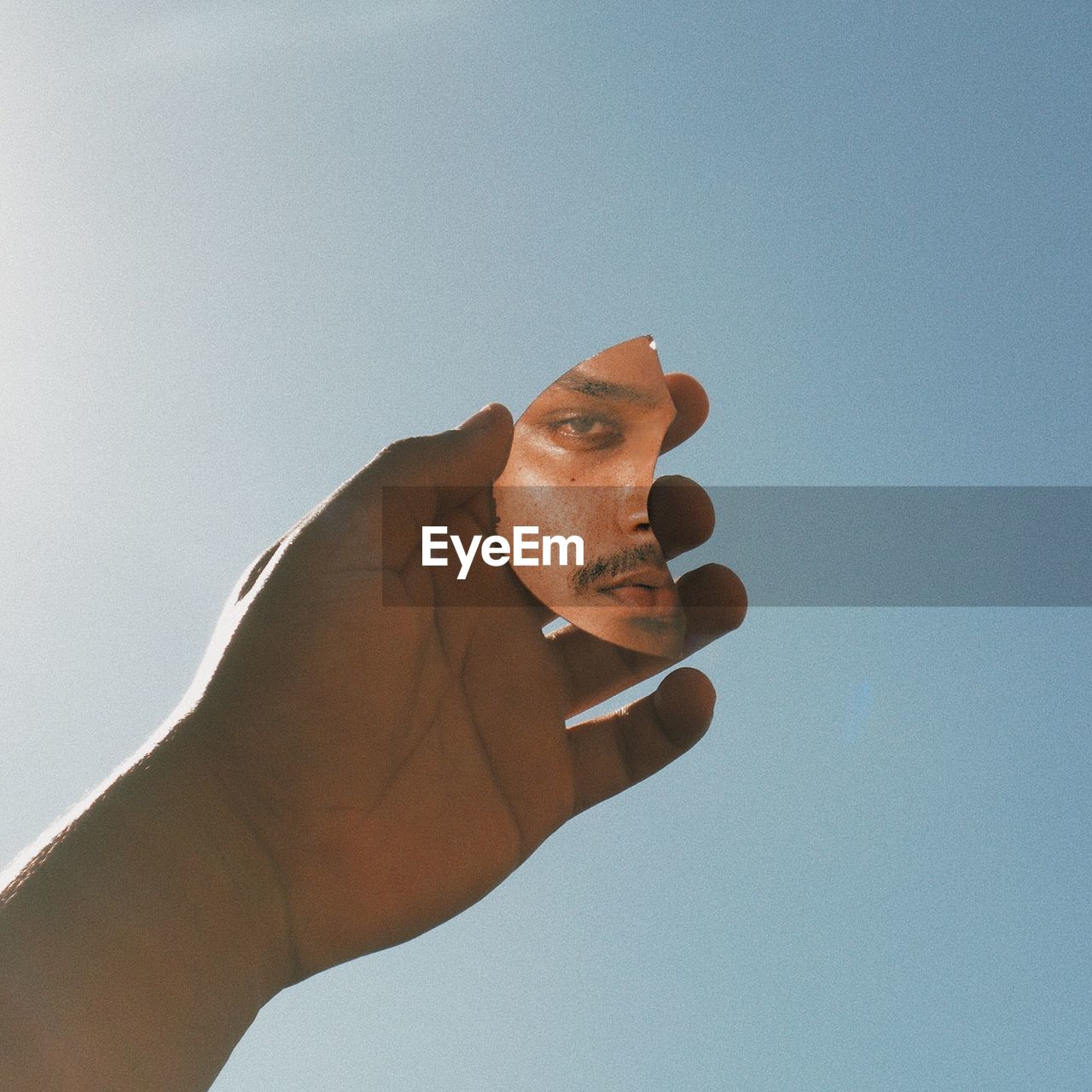 Cropped hand of man holding mirror with his reflection against clear sky