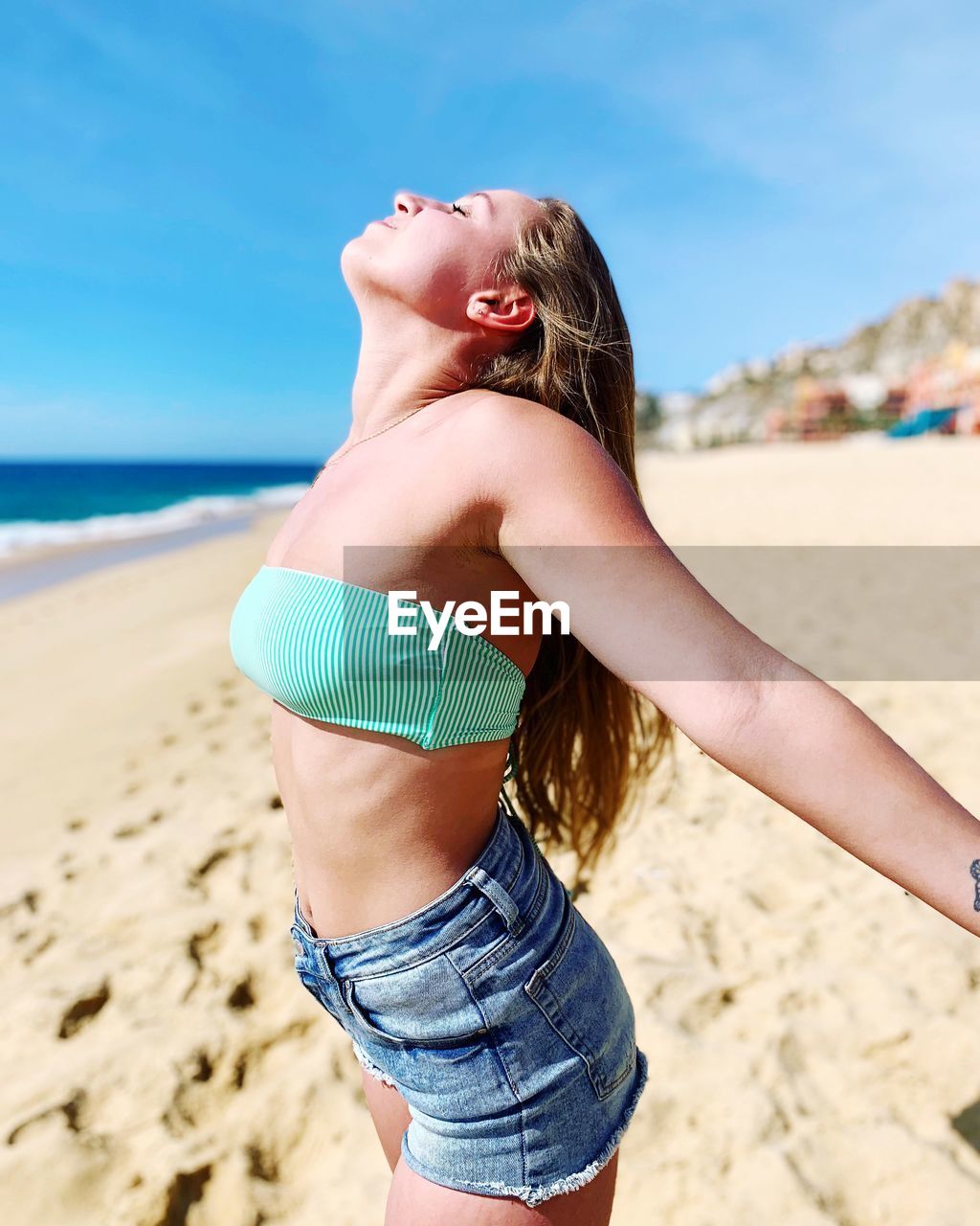 Side view of young woman with arms outstretched standing at beach against blue sky