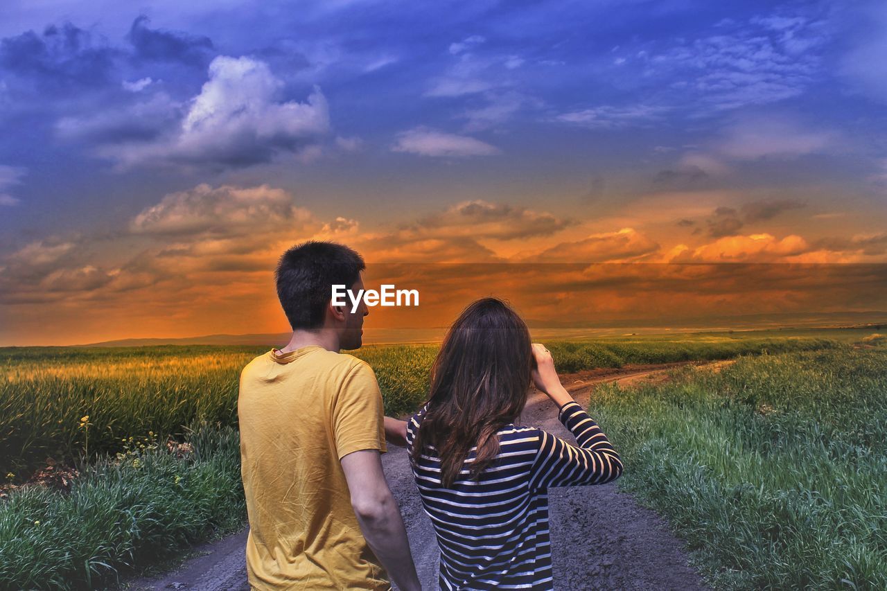 Couple on road amidst field against sky during sunset