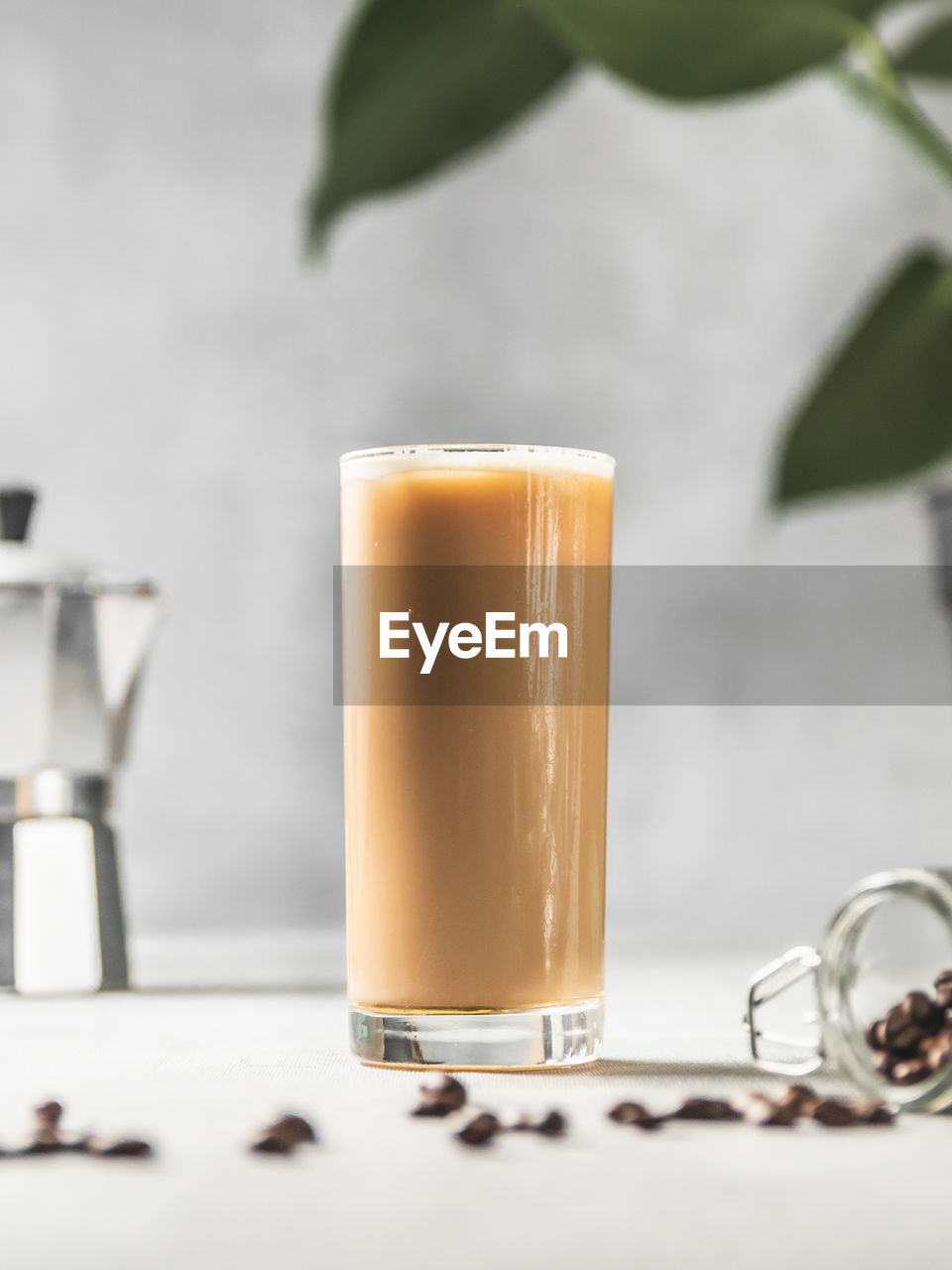 Close-up of iced coffee on table with moka pot, monstera plant, coffee beans against concrete wall