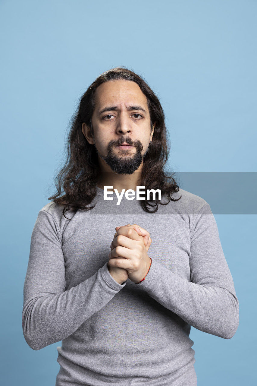 studio shot, one person, colored background, portrait, beard, adult, facial hair, blue background, gray, blue, waist up, looking at camera, front view, indoors, gray background, men, casual clothing, standing, photo shoot, brown hair, hairstyle, sitting, emotion, young adult, person, long hair, hand, looking, individuality, clothing, finger, serious