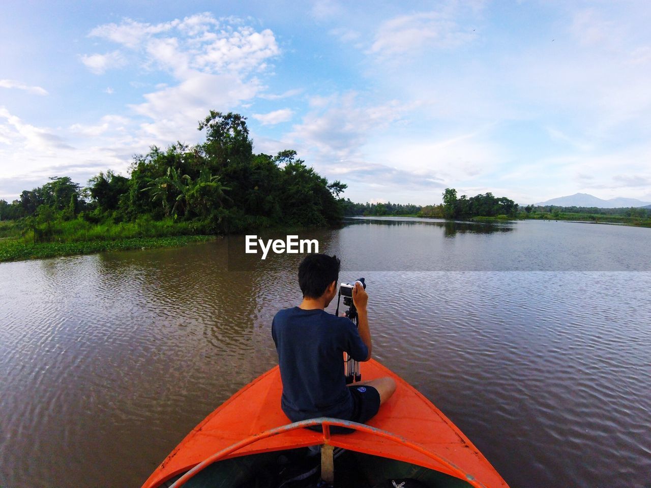 REAR VIEW OF MAN SITTING BY LAKE AGAINST SKY