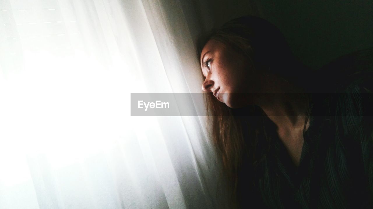 Sad woman looking away while sitting by window curtain at home