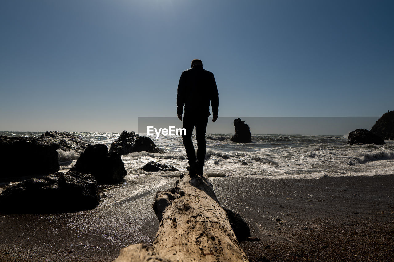 Person walking on large driftwood log toward waves at edge of beach