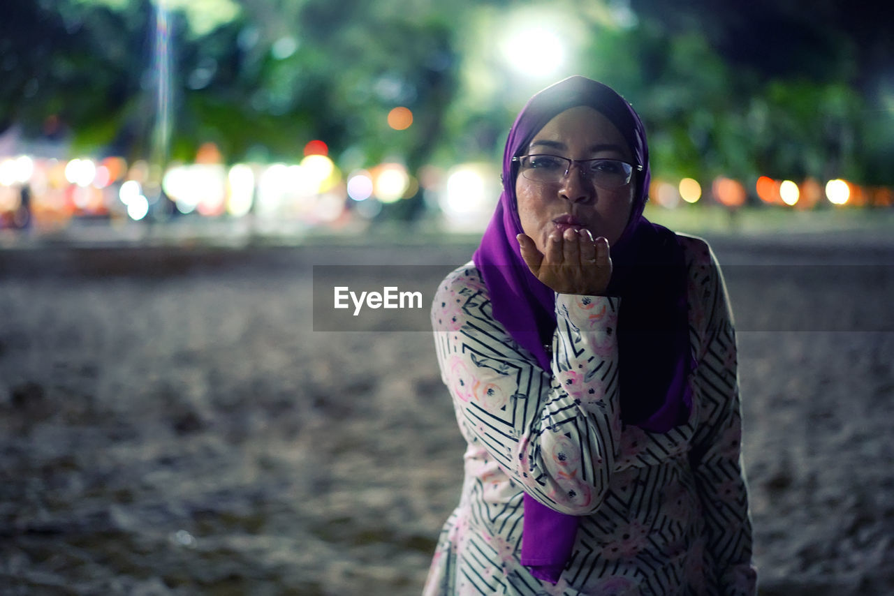 Portrait of mid adult woman blowing kiss while standing on beach at night