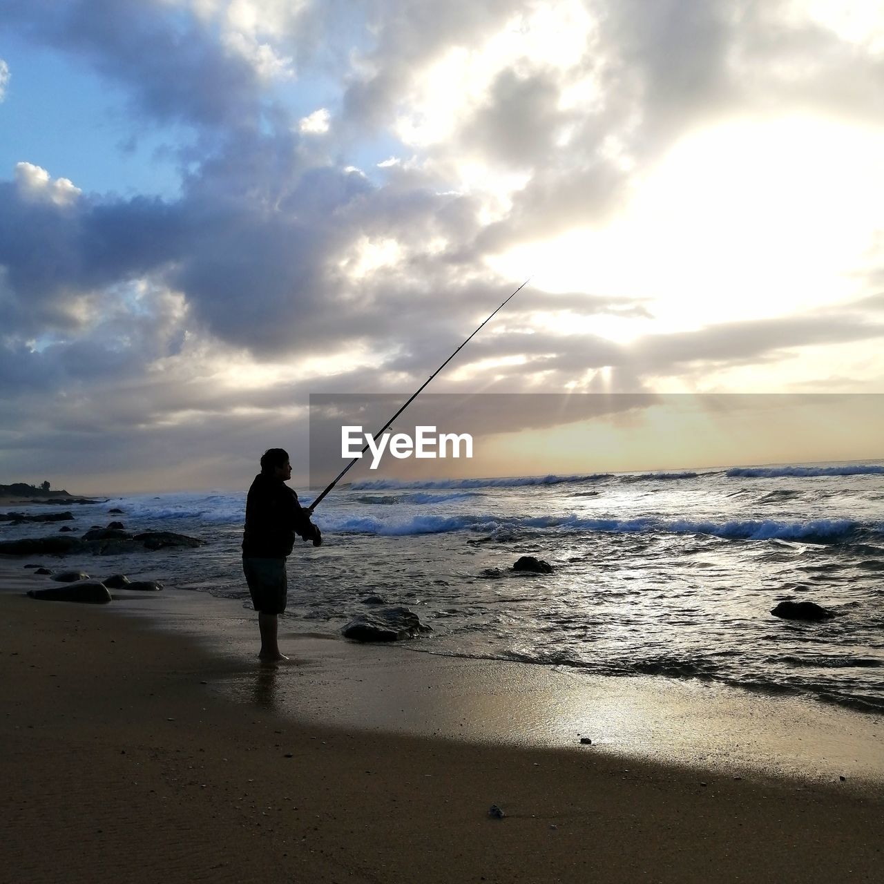Man fishing at beach against sky during sunset