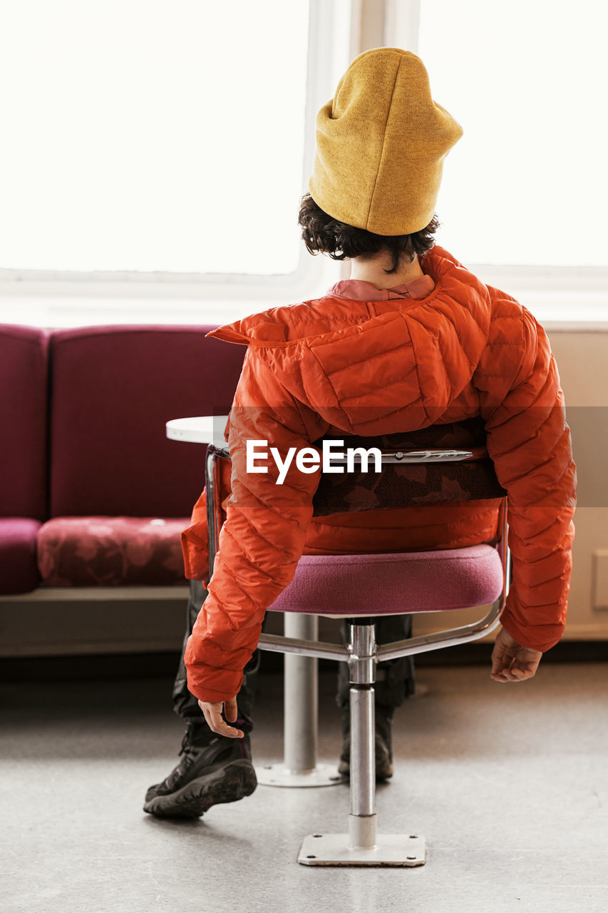 Back view of anonymous female in orange coat and yellow hat chilling on chair against sofa and window in sunlit room