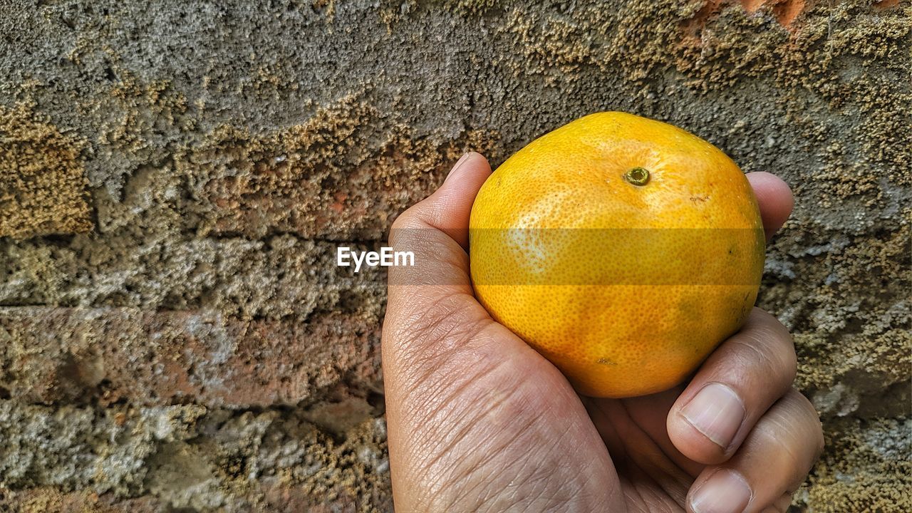 CLOSE-UP OF PERSON HAND HOLDING YELLOW FRUIT ON WALL