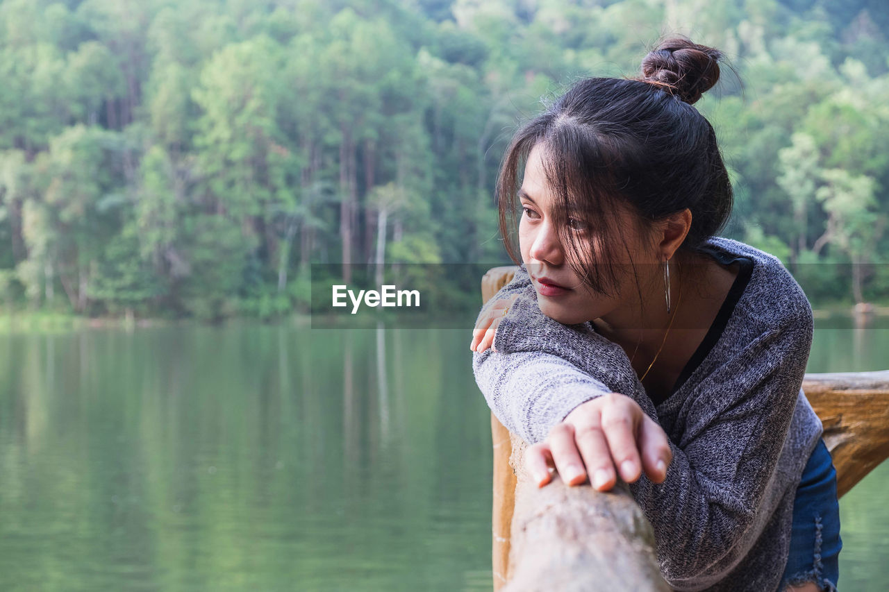 Thoughtful woman standing by railing against lake in forest