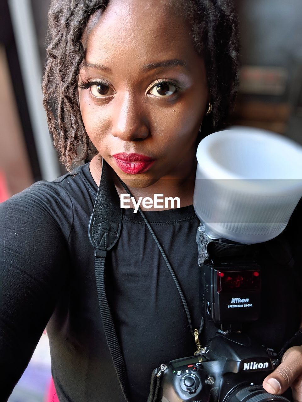 PORTRAIT OF YOUNG WOMAN HOLDING CAMERA WHILE STANDING IN OFFICE