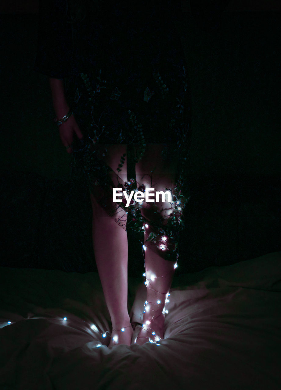 Low section of woman with illuminated string lights standing on bed in darkroom