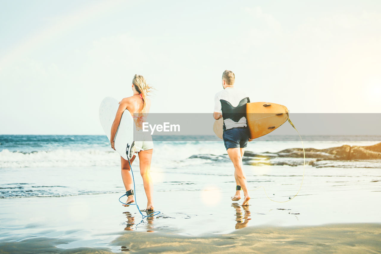 Rear view of friends with surfboards walking towards sea on shore at beach against sky