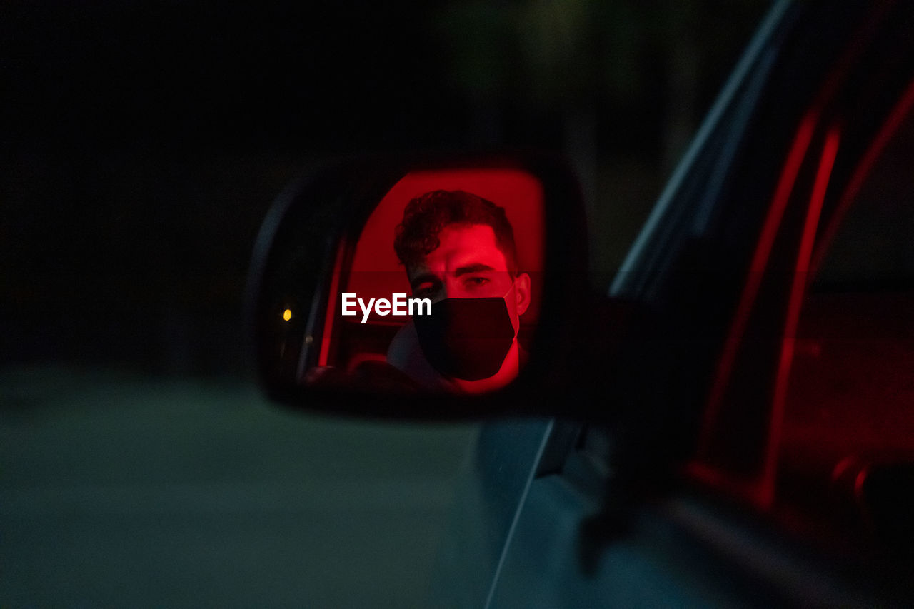 Anonymous serious male driver in face mask sitting in modern car with red neon light parked on street at night and looking in side mirror during pandemic