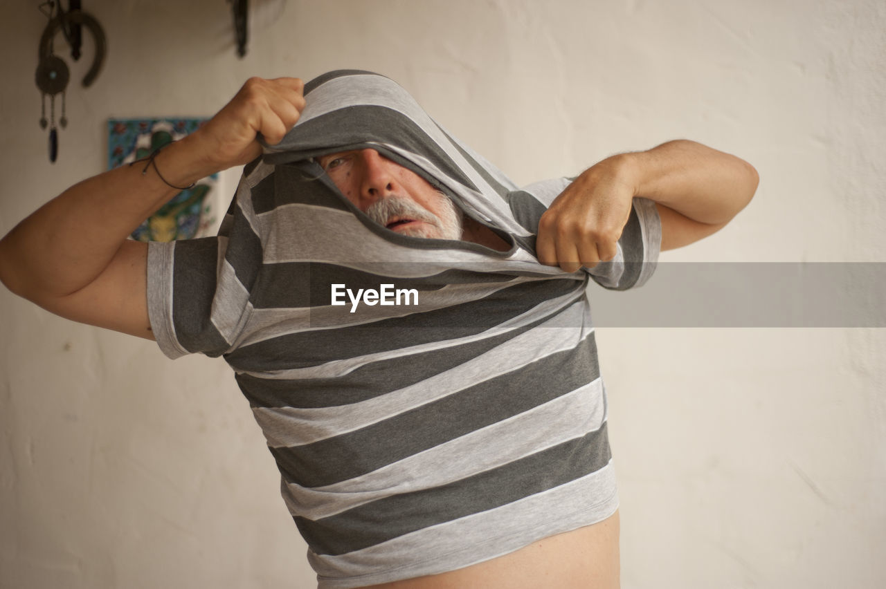 Man removing t-shirt while standing against wall at home