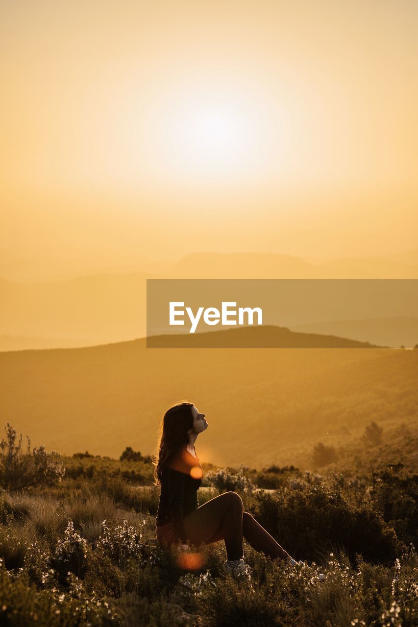 Side view of peaceful female traveler sitting on hill with closed eyes and enjoying nature in highlands at sundown