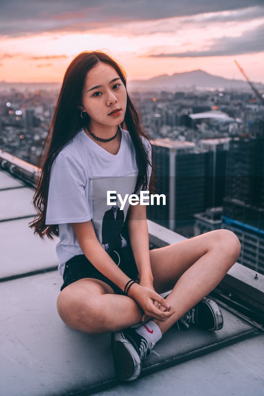 PORTRAIT OF YOUNG WOMAN SITTING ON CITY AGAINST SKY