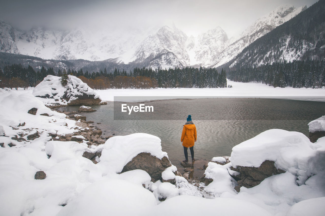 Rear view of woman standing at lakeshore against snowcapped mountains