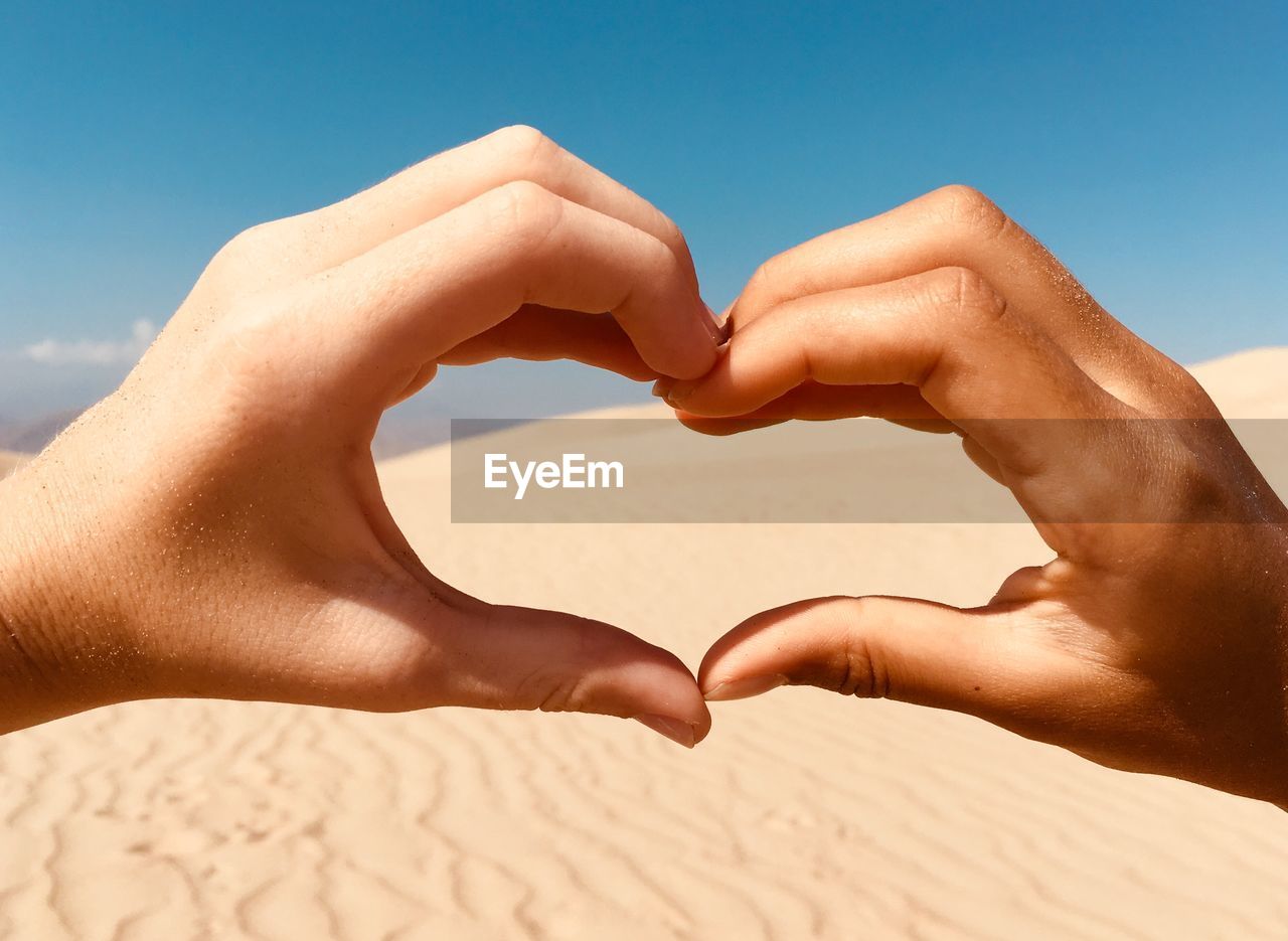 Cropped hands of people making heart shape against sand and sky