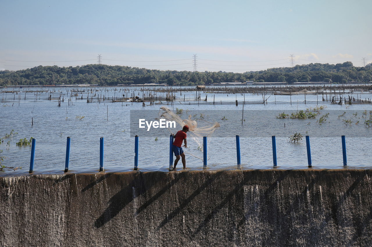 MAN FISHING AT RIVERBANK AGAINST SKY