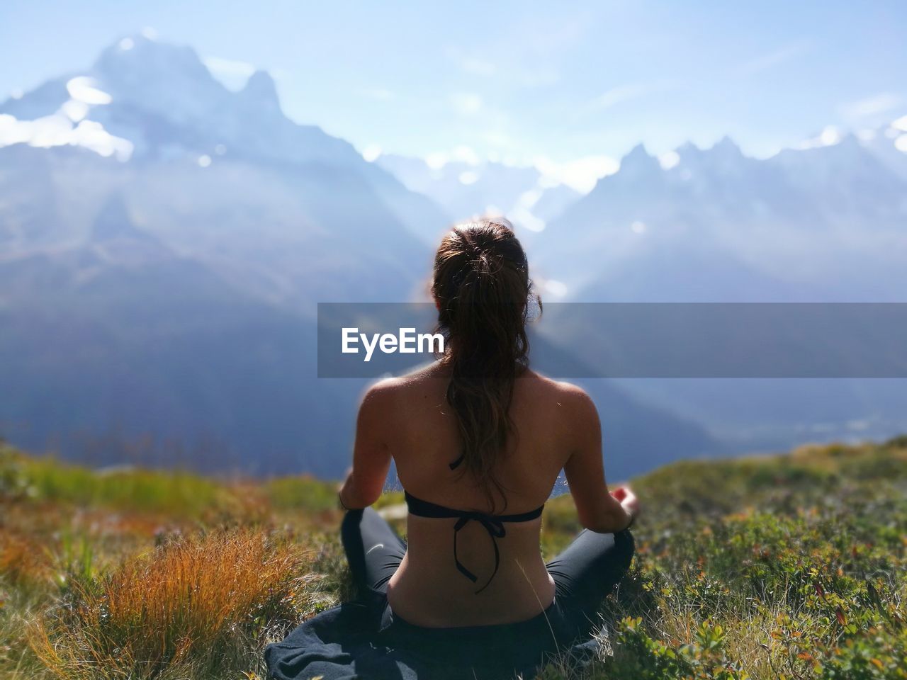 Rear view of woman meditating while sitting on grassy field against sky