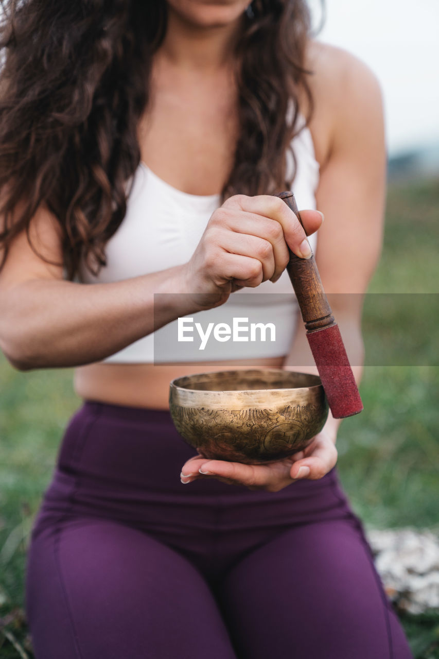 Unrecognizable female athlete in sportswear holding bell and meditating during yoga session in nature