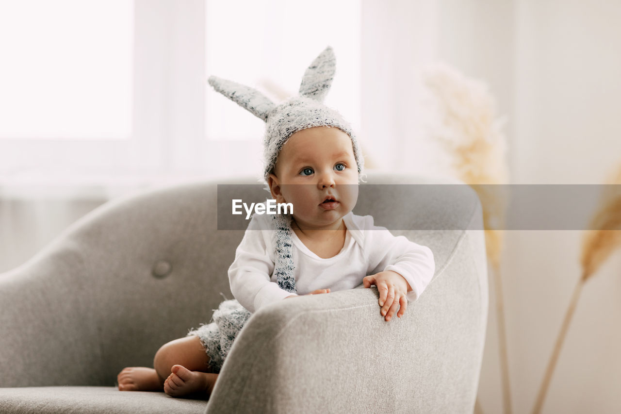 Cute baby wearing bunny cap looking away while sitting on sofa