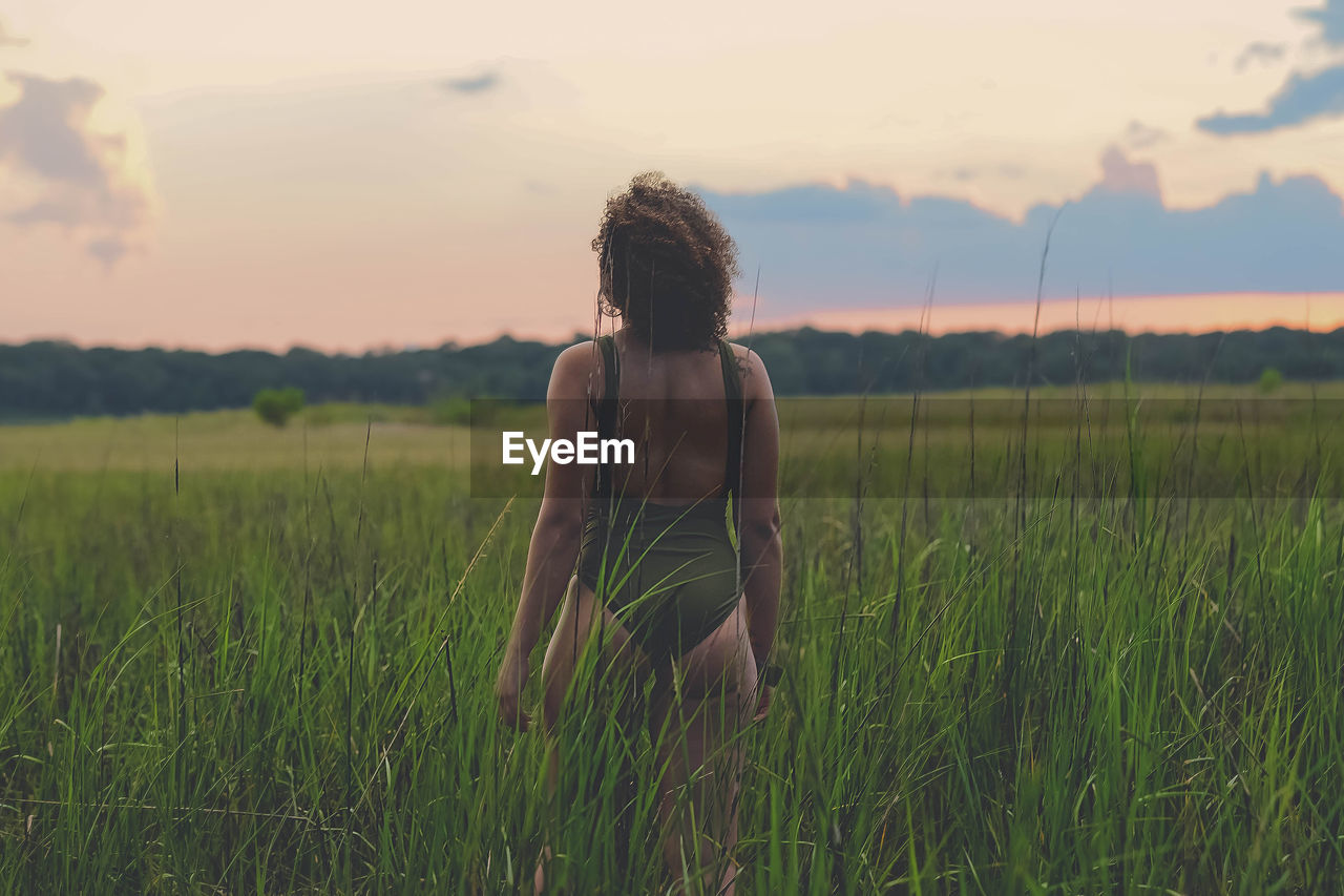 Rear view of young woman in swimwear standing on field against sky during sunset