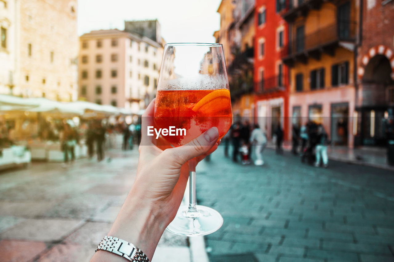 Female hand with glass of orange cocktail spritz near old buildings. sunny day in verona, italy