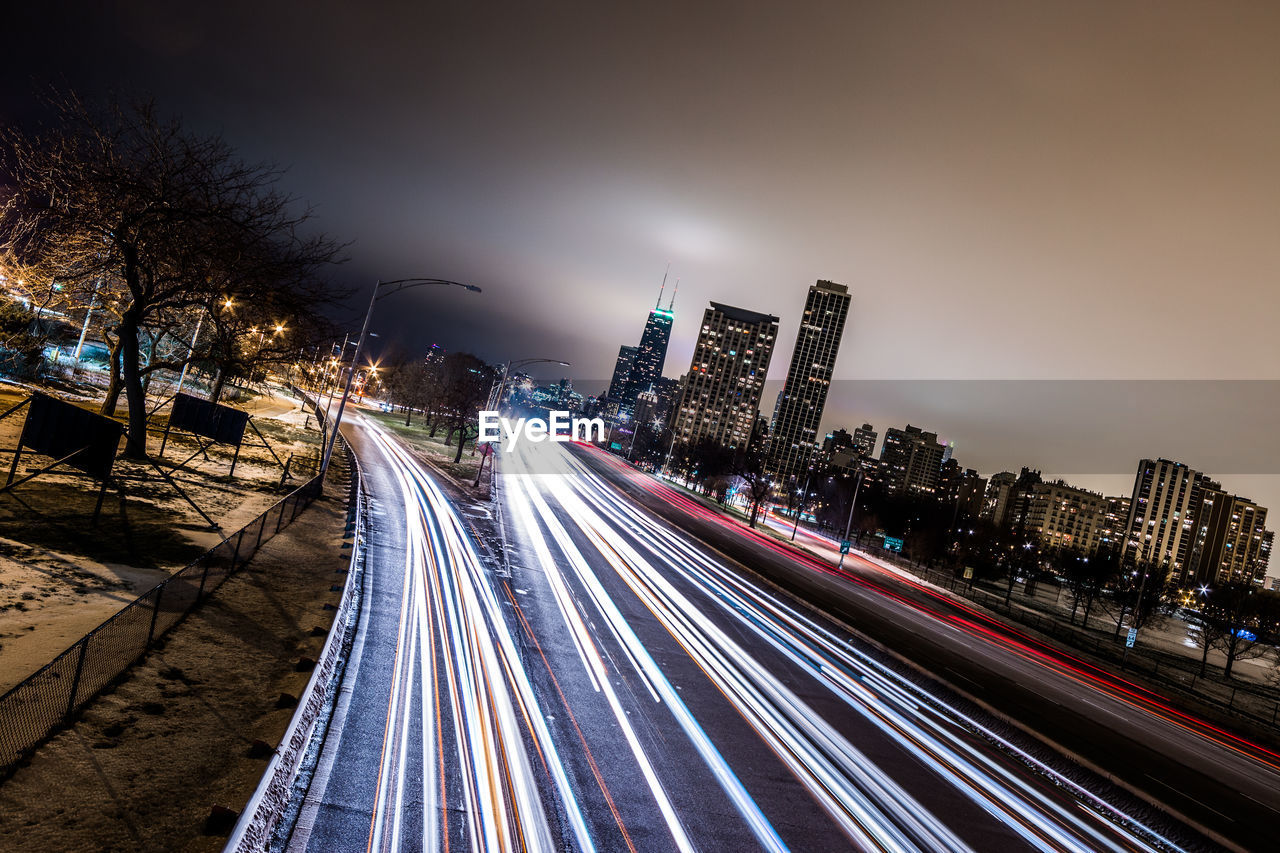 Light trails on city street by modern buildings against cloudy sky at night