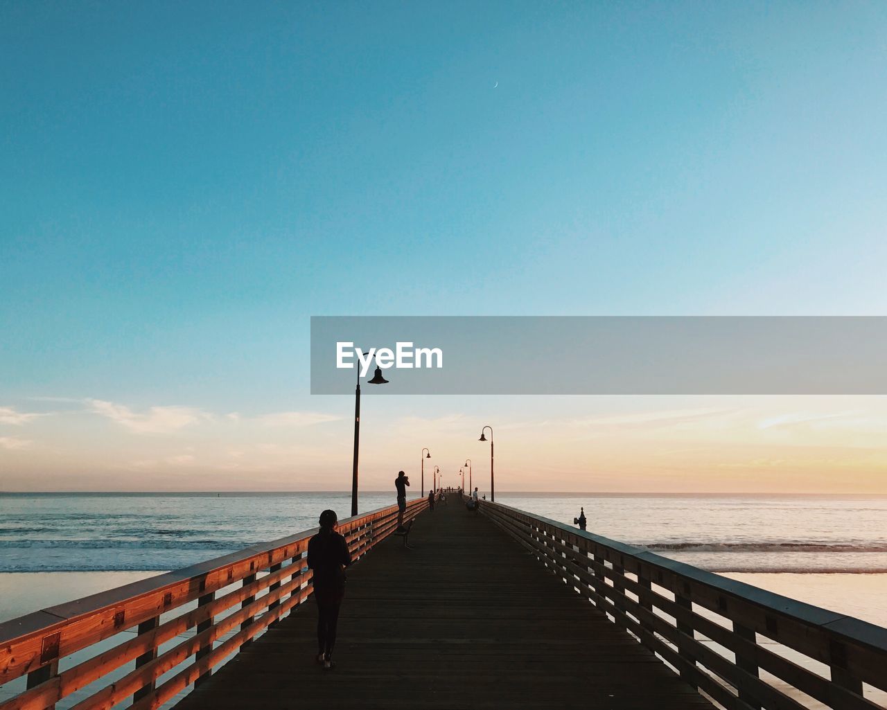 Rear view of silhouette woman walking on pier at beach against blue sky during sunset