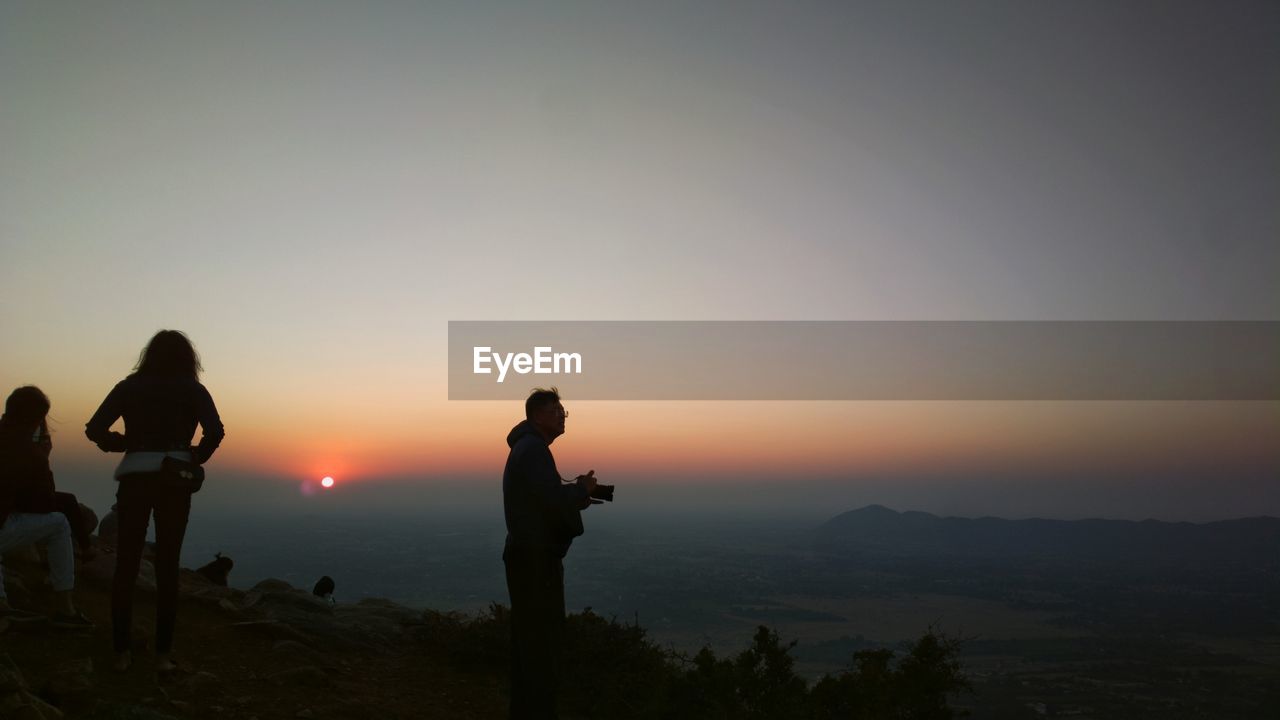 SILHOUETTE MAN PHOTOGRAPHING AT SUNSET AGAINST SKY