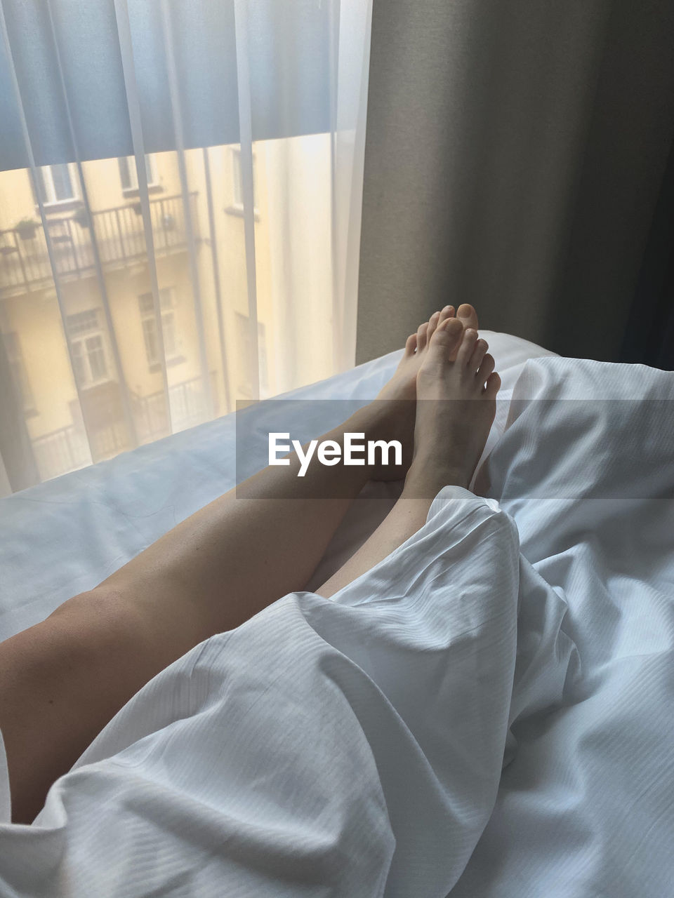 bed, indoors, bedroom, furniture, sheet, one person, domestic room, relaxation, linen, adult, textile, lifestyles, curtain, lying down, bed sheet, women, interior design, home interior, dress, barefoot, resting, white, domestic life, comfortable, high angle view, pillow, morning, day, hotel, human leg