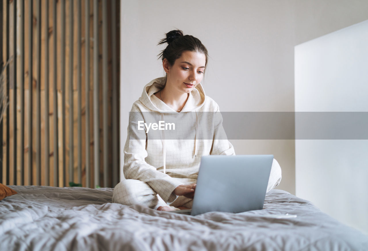 Beautiful smiling woman student freelancer with dark long hair in casual using laptop on bed at home