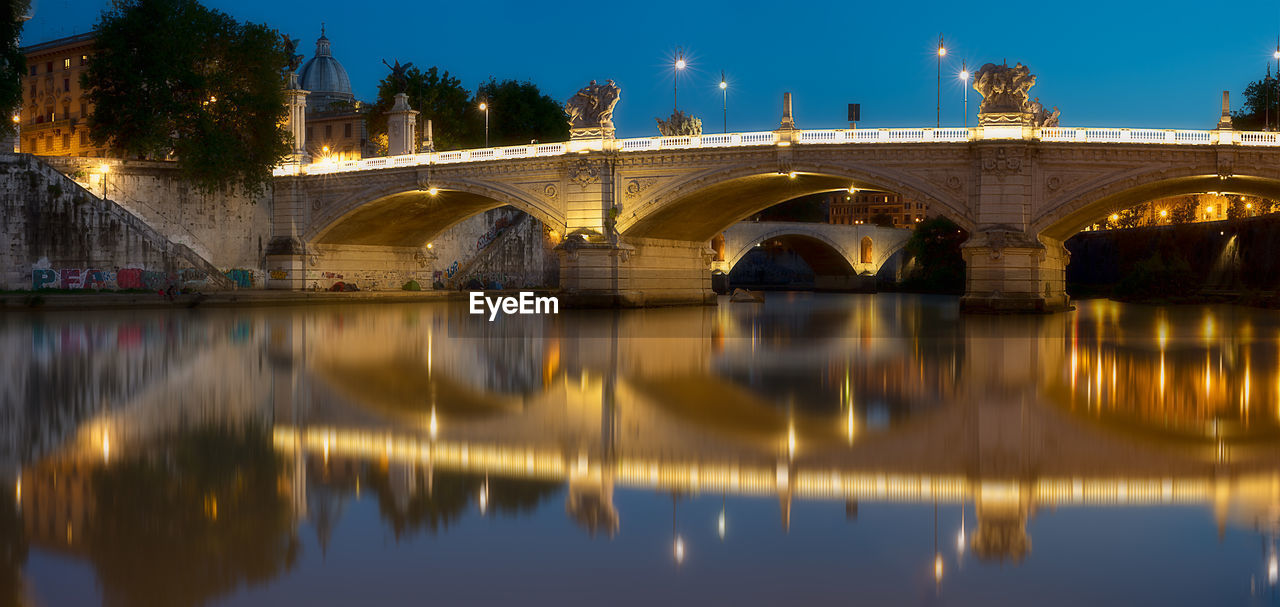 Reflection of bridge on river against sky in city at night