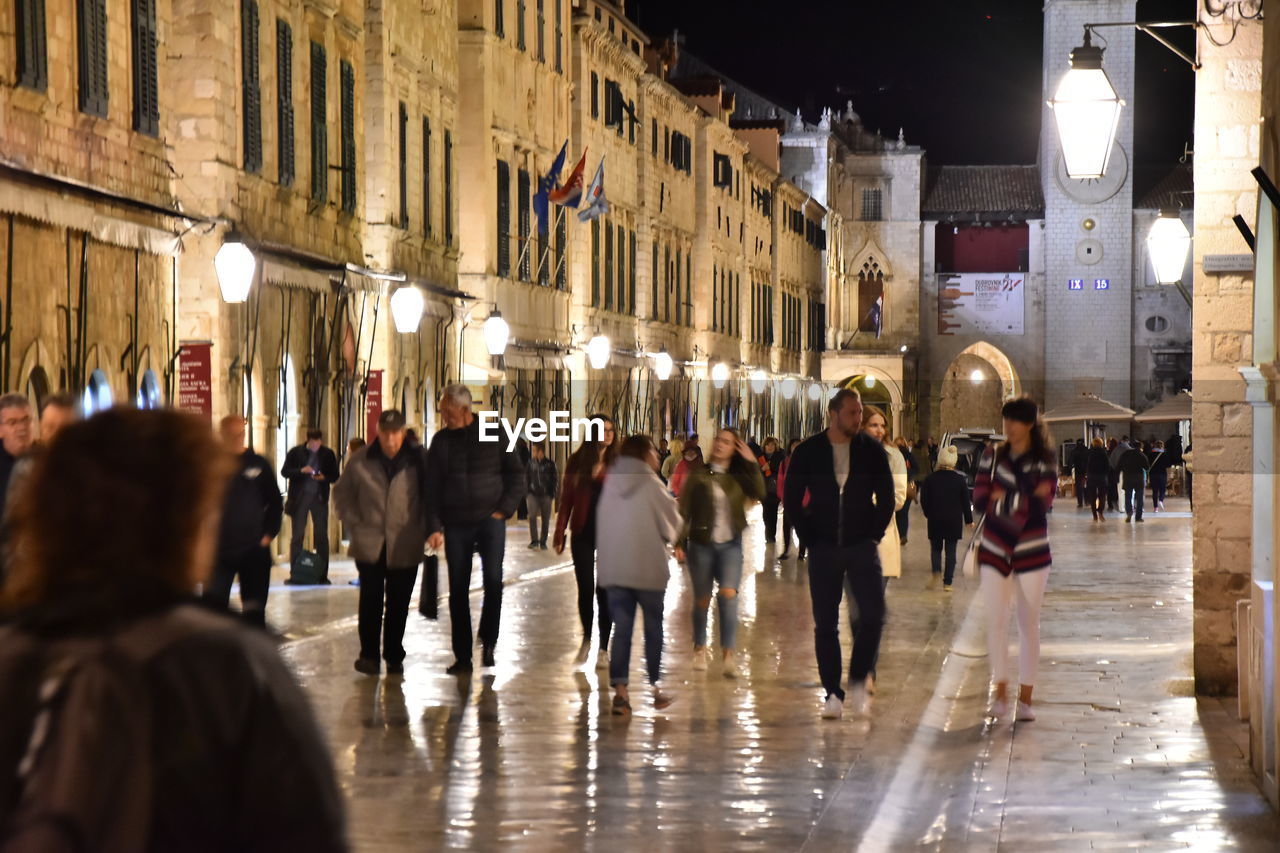 PEOPLE WALKING ON ILLUMINATED STREET IN CITY