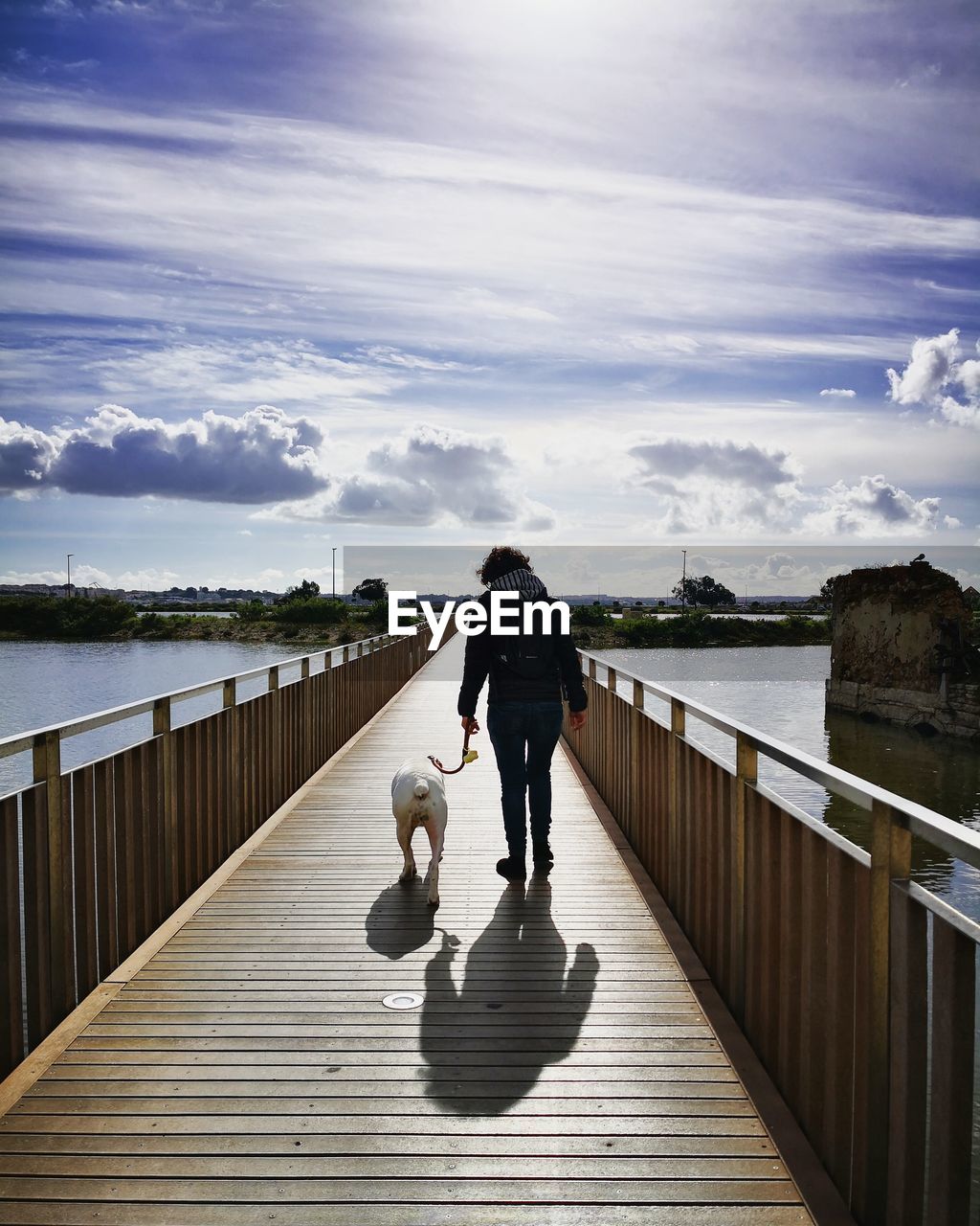 Rear view of woman with dog walking on footbridge over river against sky