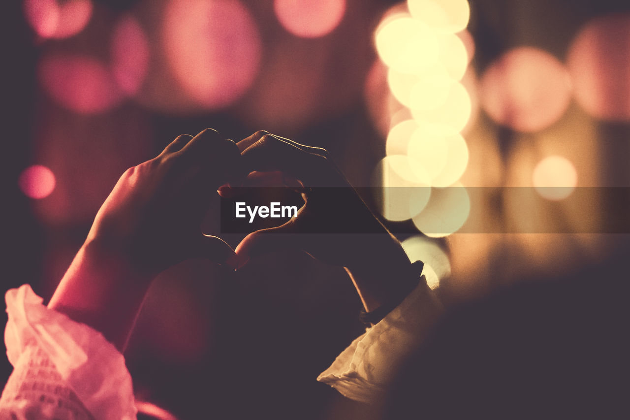 Cropped image of woman forming heart shape against lens flare at night