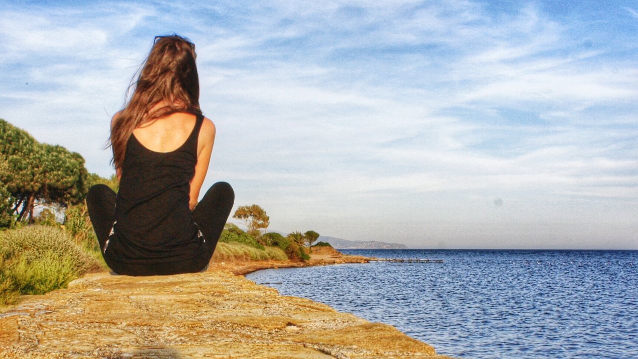 Rear view of woman sitting at seaside against cloudy sky