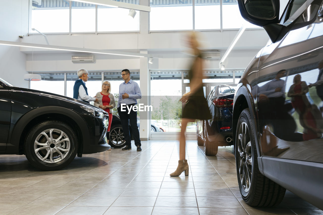 Car dealer advising customers with woman walking in foreground