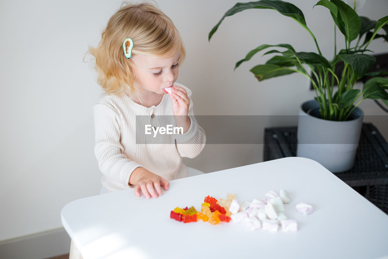 Adorable cute caucasian blonde curly-haired baby girl two, three year old eating jelly, sweet gummy